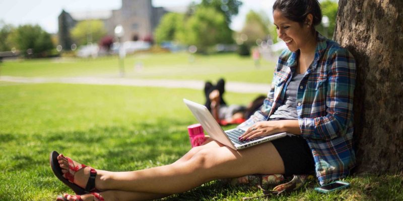 Female student sitting under the shade of a tree on the drillfield typing on a laptop.