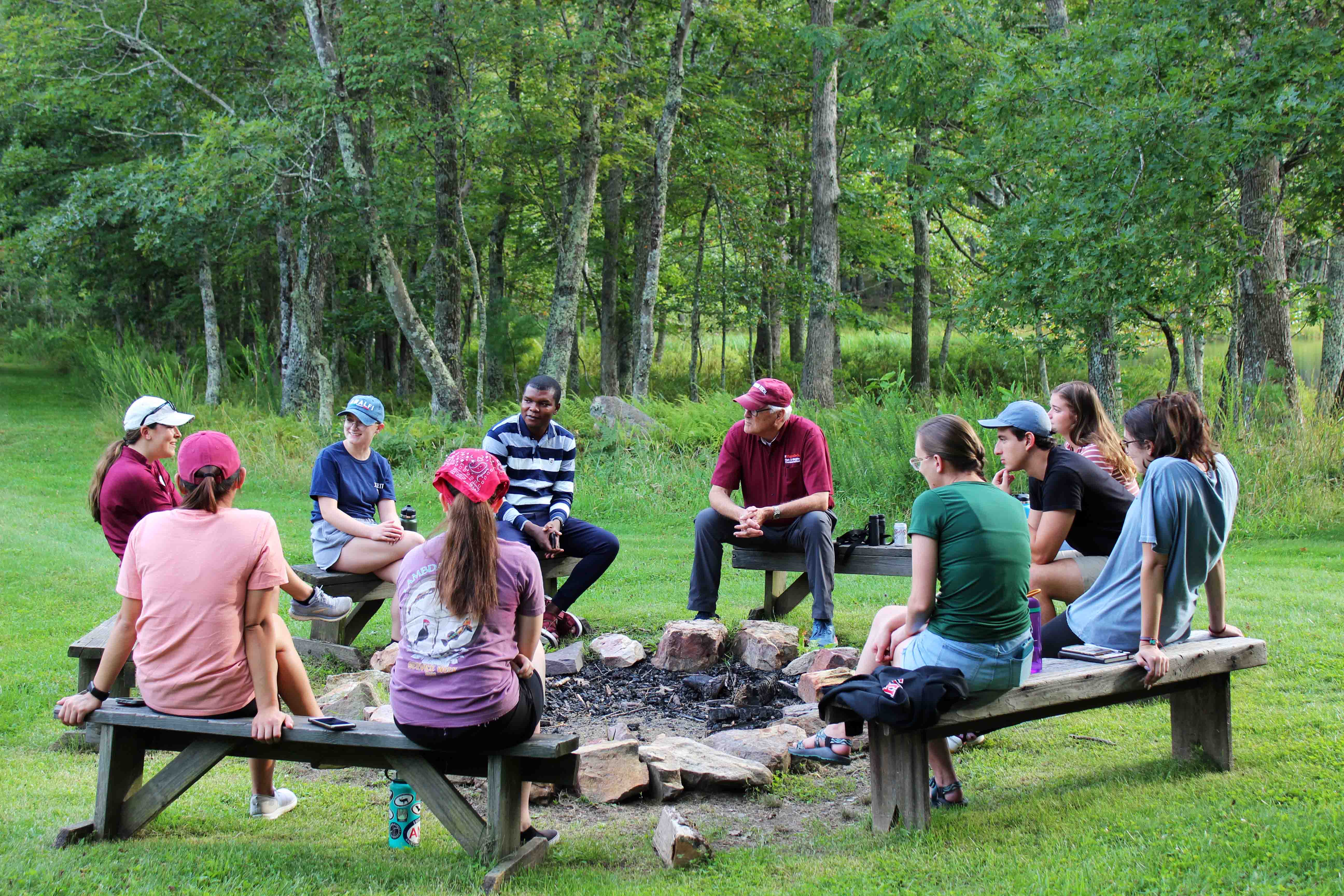 Students and a professor sit on benches in a circle around a campfire.